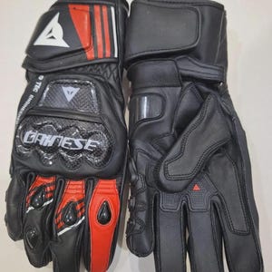 May include: Pair of black motorcycle gloves with red and white accents. The gloves feature the brand name "DAINESE" and include protective padding on the knuckles and wrist. The gloves are made of leather and have a secure wrist strap.