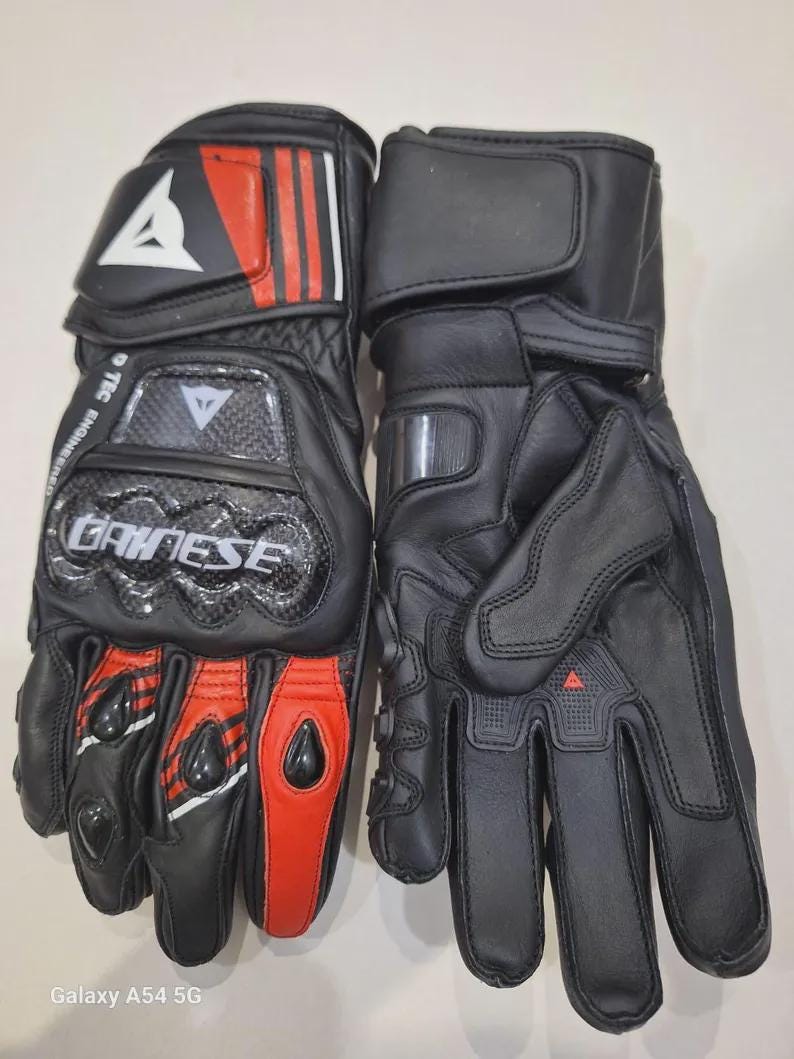 May include: Pair of black motorcycle gloves with red and white accents. The gloves feature the brand name "DAINESE" and include protective padding on the knuckles and wrist. The gloves are made of leather and have a secure wrist strap.