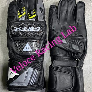 May include: Pair of black leather motorcycle gloves with carbon fiber knuckle protection. Features gray and yellow accents, the brand name "DAINESE", and the "Veloce Racing Lab" logo.
