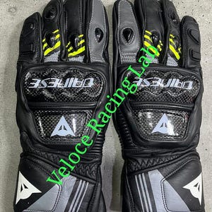 May include: Pair of black motorcycle gloves with carbon fiber knuckle protection. The gloves feature gray and yellow accents, the brand name "DAINESE", and a white "A" logo.