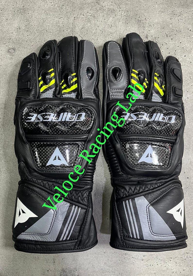 May include: Pair of black motorcycle gloves with carbon fiber knuckle protection. The gloves feature gray and yellow accents, the brand name "DAINESE", and a white "A" logo.