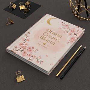 Puede incluir: Un cuaderno de tapa dura en color crema con ramas de cerezo en flor de color rosa, una luna creciente dorada y el texto "Dream. Create. Bloom." con "MonivikaDigital" impreso debajo. El cuaderno está acompañado por dos bolígrafos.