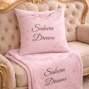 Puede incluir: Un juego de manta y cojín rosas con las palabras "Sakura Dreams" impresas. El juego presenta un diseño floral con ramas de flores de cerezo y pétalos que caen. Los artículos se exhiben en un sofá beige ornamentado.