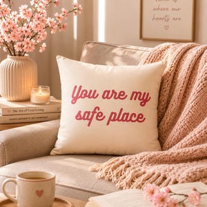 Puede incluir: Un cojín crema con el texto "You are my safe place" en escritura roja está sobre un sillón beige. Una manta rosa, una taza con un corazón y un jarrón con flores rosas completan la escena.