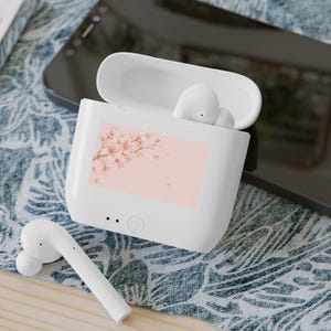 Puede incluir: Auriculares inalámbricos blancos y estuche de carga. El estuche tiene un fondo rosa con un diseño floral. Un auricular está fuera del estuche y dos dentro. El estuche está abierto y los auriculares son blancos.