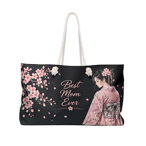 Puede incluir: Bolso tote negro con asas de cuerda blancas. El bolso presenta un diseño floral con flores rosas y el texto "Best Mom Ever". Una mujer con un kimono rosa está en el lado derecho del bolso.