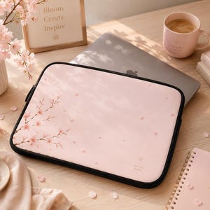 Puede incluir: Una funda rectangular para portátil en un tono rosa pastel suave, con una ilustración de una rama de cerezo en el lado izquierdo y pétalos dispersos por la superficie. La funda tiene un borde con cremallera negra y un pequeño logotipo.