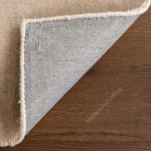Puede incluir: Una esquina de una alfombra beige con una superficie texturizada visible y un fleco blanco. La parte inferior de la alfombra es gris y blanca, revelando la construcci&oacute;n tejida. La alfombra est&aacute; sobre una superficie de madera marr&oacute;n.