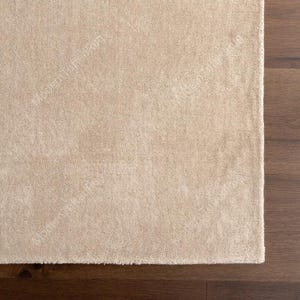 Puede incluir: Una alfombra rectangular beige con una superficie suave y texturizada. La alfombra est&aacute; colocada junto a un suelo de madera marr&oacute;n oscuro. Se ve la esquina de la alfombra, mostrando su borde y textura. La alfombra parece estar hecha de un material afelpado.