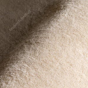Puede incluir: Primer plano de una alfombra beige con mechones. La alfombra tiene una superficie suave y texturizada con una pila densa. Las palabras "ModernTuftedRug" est&aacute;n impresas en diagonal en la esquina superior izquierda. La alfombra parece estar hecha de un material afelpado.