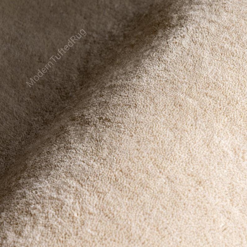 Puede incluir: Primer plano de una alfombra beige con mechones. La alfombra tiene una superficie suave y texturizada con una pila densa. Las palabras "ModernTuftedRug" est&aacute;n impresas en diagonal en la esquina superior izquierda. La alfombra parece estar hecha de un material afelpado.