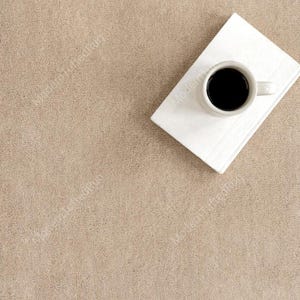 Puede incluir: Vista a&eacute;rea de una alfombra beige con un libro blanco y una taza de caf&eacute; blanca llena de caf&eacute; negro. La alfombra tiene una superficie texturizada. La taza est&aacute; sobre el libro.