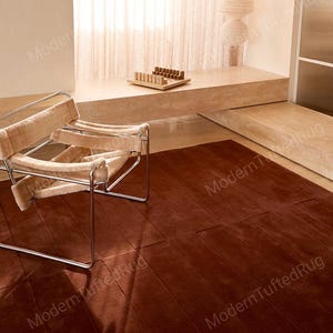 May include: A large, rectangular, rust-coloured rug with a tufted design. The rug is placed on a wooden floor, and a modern chair with a metal frame and beige fabric is in the foreground. The room has a minimalist design with natural light.