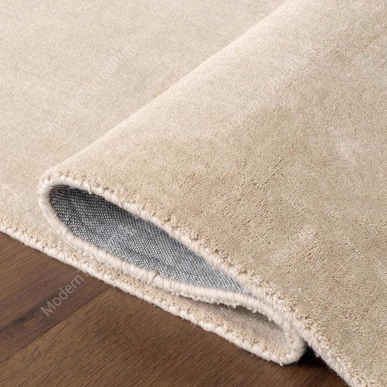 Puede incluir: Una alfombra beige enrollada con una superficie suave y texturizada. La parte inferior de la alfombra es de tela gris. La alfombra est&aacute; sobre un suelo de madera marr&oacute;n oscuro.