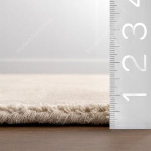 Puede incluir: Primer plano de una alfombra beige con superficie texturizada. El borde de la alfombra es visible, mostrando un pelo grueso. Una regla se coloca junto a la alfombra, con medidas de hasta 8 cm.