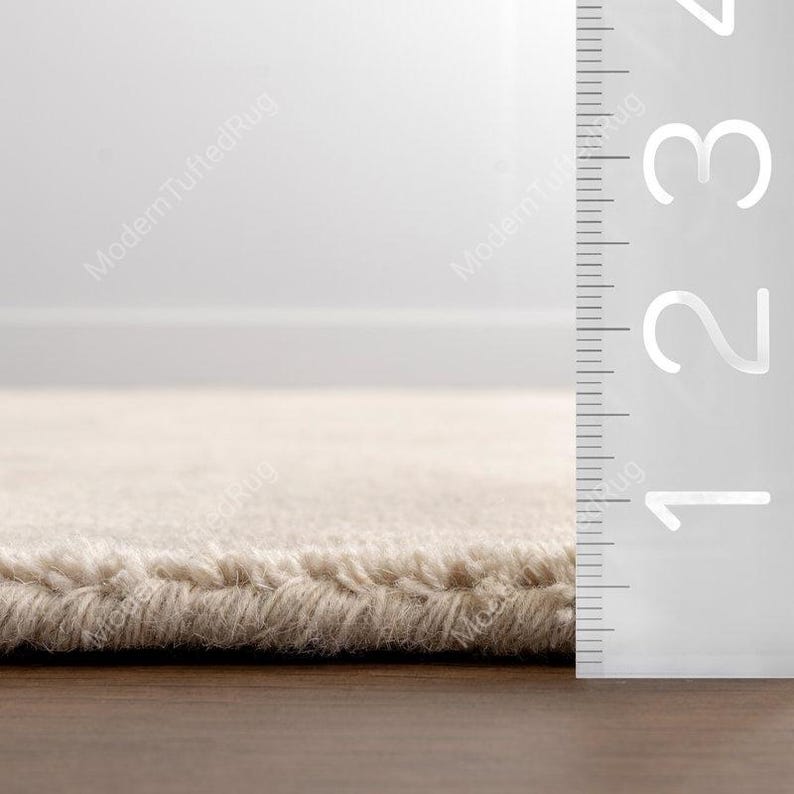 Puede incluir: Primer plano de una alfombra beige con superficie texturizada. El borde de la alfombra es visible, mostrando un pelo grueso. Una regla se coloca junto a la alfombra, con medidas de hasta 8 cm.