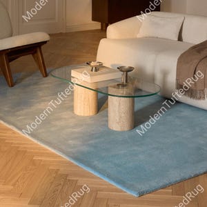 Könnte beinhalten: Ein hellblauer rechteckiger Teppich mit Farbverlauf, auf einem Fischgrät-Holzboden. Ein Couchtisch mit Glasplatte und zylindrischen Steinsockeln steht auf dem Teppich. Ein weißes Sofa und ein Stuhl sind im Hintergrund.
