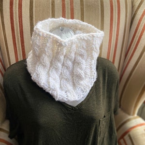 May include: White knitted neck warmer with a cable knit pattern. The neck warmer is displayed on a brown and white striped armchair.