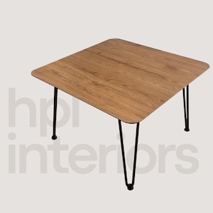 Puede incluir: Una mesa cuadrada con una superficie de grano de madera marrón claro y esquinas redondeadas. La mesa está sostenida por cuatro patas delgadas de metal negro en forma de horquilla que se inclinan hacia afuera. El texto hpi interiors es visible.