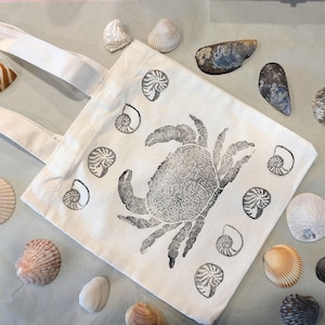 May include: A white canvas tote bag with a large black crab illustration and smaller nautilus shell designs. Various seashells in shades of white, tan, and brown are scattered around the bag, creating a coastal theme.