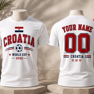 Op de afbeelding: Wit t-shirt met "CROATIA" in rood en blauw, een voetbalafbeelding en de tekst "WORLD CUP 2026". De achterkant toont "YOUR NAME", het nummer "00" en "CROATIA".