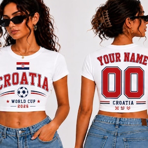 K&ouml;nnte beinhalten: Wei&szlig;es Crop-Top mit "CROATIA" in roter und blauer Schrift, einer Fu&szlig;ballgrafik und "WORLD CUP 2026". Auf der R&uuml;ckseite stehen "YOUR NAME" und die Nummer "00" in Rot.
