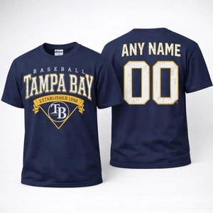 Könnte beinhalten: Marineblaues Baseball-T-Shirt mit dem Text "TAMPA BAY" in Weiß und Gold, gegründet 1998. Auf der Rückseite des Shirts steht "ANY NAME" über der Nummer "00" in Weiß und Gold.