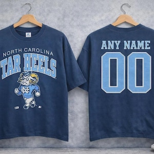 May include: Navy blue t-shirt featuring "North Carolina Tar Heels" text and a ram mascot graphic on the front. The back displays "ANY NAME" and the number "00" in light blue.