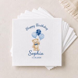 May include: A stack of white paper napkins featuring a printed illustration of a light brown teddy bear wearing a blue bow tie and holding three blue balloons. The text reads Happy Birthday, the name Sophia, and the date 11.10.2026 in blue ink.
