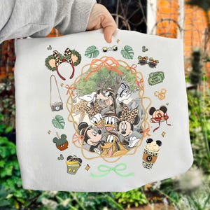 Puede incluir: Bolso tote blanco con un diseño temático de Disney. La imagen presenta a Mickey y Minnie Mouse, el Pato Donald y Goofy, junto con elementos de estilo safari como lazos y gafas de sol con estampado animal, e iconos relacionados con el parque.
