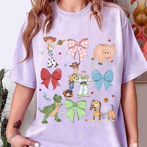 May include: A light purple t-shirt featuring colorful cartoon characters and bows. The design includes a cowgirl, a cowboy, a space ranger, a pig, a dinosaur, a dog, and several bows in various colors.