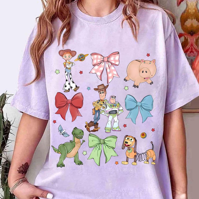 May include: A light purple t-shirt featuring colorful cartoon characters and bows. The design includes a cowgirl, a cowboy, a space ranger, a pig, a dinosaur, a dog, and several bows in various colors.