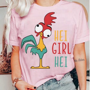May include: Pink t-shirt featuring a cartoon rooster with red, yellow, and teal accents. The text "HEI GIRL HEI" is printed in yellow, red, and teal. The shirt is made of a soft material.