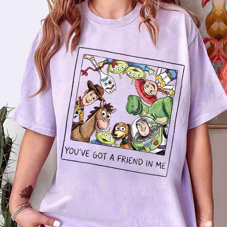 May include: A light purple t-shirt featuring a framed graphic of characters from the animated movie Toy Story. The graphic includes Woody, Buzz Lightyear, and other characters. The text "YOU'VE GOT A FRIEND IN ME" is below the graphic.