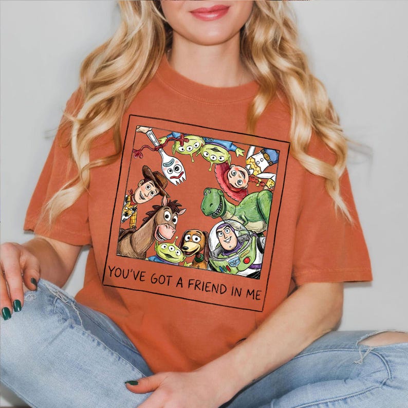 May include: Burnt orange t-shirt featuring a framed graphic of Toy Story characters, including Woody, Buzz Lightyear, and the Little Green Men. The text "YOU'VE GOT A FRIEND IN ME" is printed below the graphic. The shirt is worn by a person.