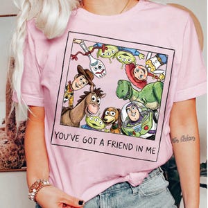 May include: Pink t-shirt featuring a framed graphic of Toy Story characters, including Woody, Buzz Lightyear, and Rex. The phrase "YOU'VE GOT A FRIEND IN ME" is printed below the image. The shirt has short sleeves and a crew neck.