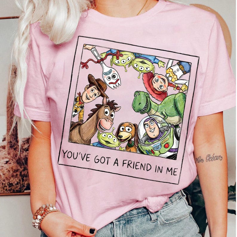 May include: Pink t-shirt featuring a framed graphic of Toy Story characters, including Woody, Buzz Lightyear, and Rex. The phrase "YOU'VE GOT A FRIEND IN ME" is printed below the image. The shirt has short sleeves and a crew neck.