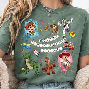 May include: Green t-shirt featuring colorful cartoon characters and the phrase "You've Got a Friend in Me." The design includes images of Woody, Buzz Lightyear, and other characters from the animated movie.