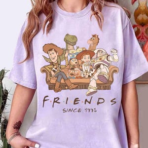 May include: A light purple t-shirt featuring a graphic of characters from the Toy Story movie franchise. The characters are seated on a brown couch. The word "FRIENDS" is printed below the graphic, with "SINCE 1995" underneath.