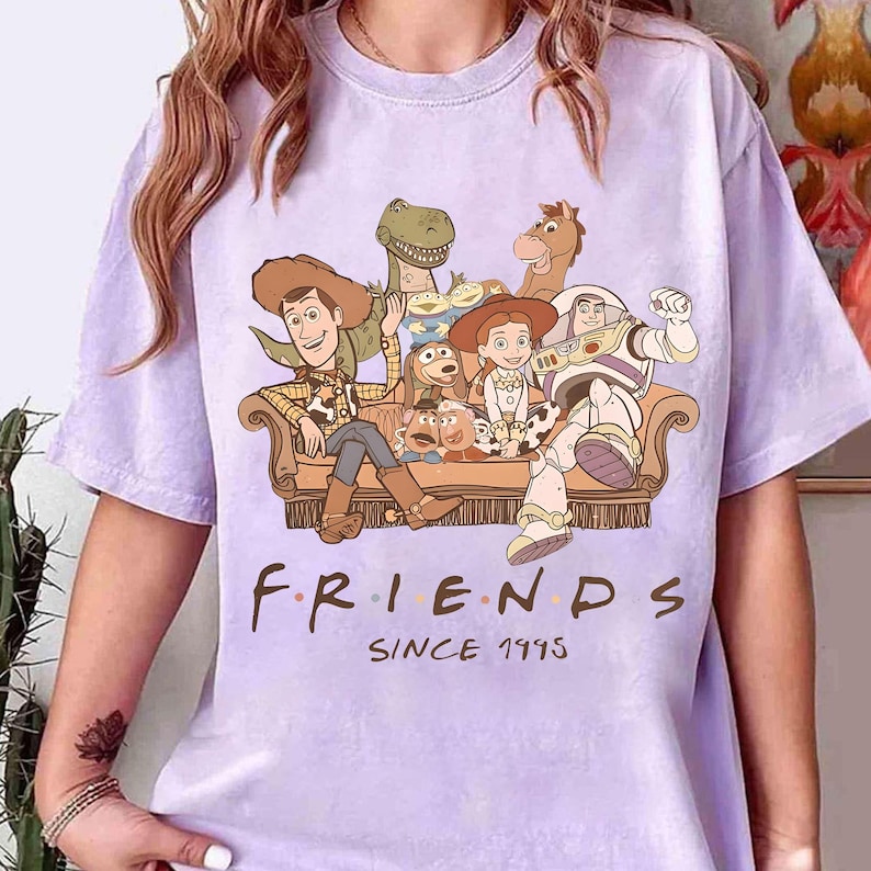 May include: A light purple t-shirt featuring a graphic of characters from the Toy Story movie franchise. The characters are seated on a brown couch. The word "FRIENDS" is printed below the graphic, with "SINCE 1995" underneath.
