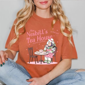 May include: Burnt orange t-shirt featuring a cartoon illustration of Buzz Lightyear in an apron, drinking tea at a table. The text "Mrs. Nesbitt's Tea House" is above the illustration, with "To Infinity and Beyond" below.