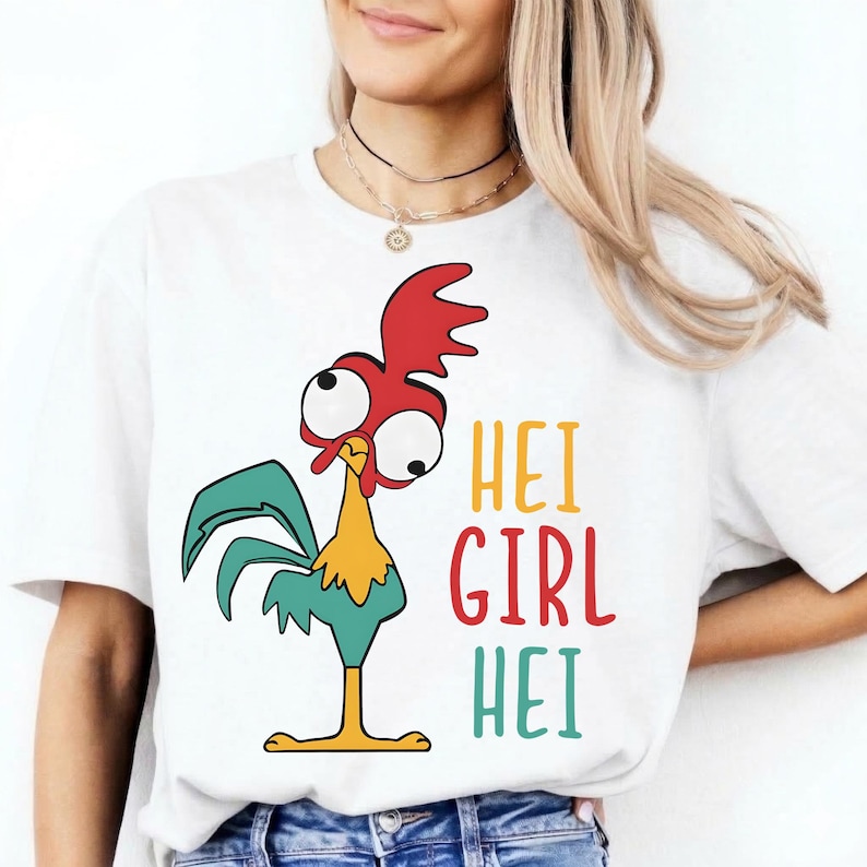 May include: White t-shirt featuring a cartoon rooster with a red comb, large eyes, and teal feathers. The text "HEI GIRL HEI" is printed in yellow, red, and teal. The shirt is a casual, everyday wear item.