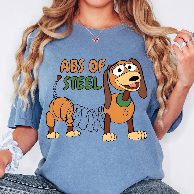 May include: A light blue t-shirt featuring a cartoon illustration of Slinky Dog with the text "ABS OF STEEL" in orange and green. The design includes the dog's body and spring-like midsection.