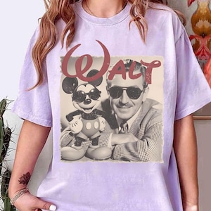 Puede incluir: Camiseta de color lila claro con un gr&aacute;fico vintage de Mickey Mouse y Walt Disney con gafas de sol. La palabra "Walt" est&aacute; impresa en rojo, con una fuente estilizada sobre la imagen. La camiseta tiene mangas cortas.