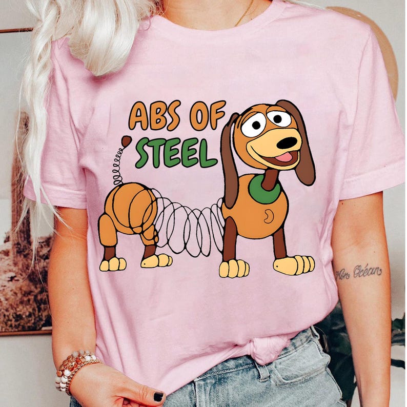 May include: Pink t-shirt featuring a cartoon image of Slinky Dog with the text "ABS OF STEEL" in orange and green. The dog is brown, tan, and yellow with a green collar. The shirt is a short-sleeved crew neck.