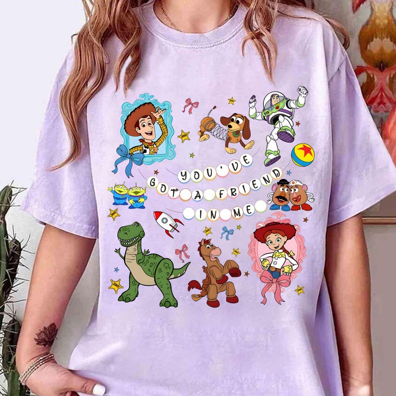 May include: A light purple t-shirt featuring colorful cartoon characters and the text "YOU'VE GOT A FRIEND IN ME". The design includes Woody, Buzz Lightyear, and other characters from the Toy Story franchise.