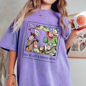 May include: A purple t-shirt featuring a framed graphic of Toy Story characters, including Woody, Buzz Lightyear, and Rex. The text "YOU'VE GOT A FRIEND IN ME" is printed below the graphic. The shirt is a solid color.