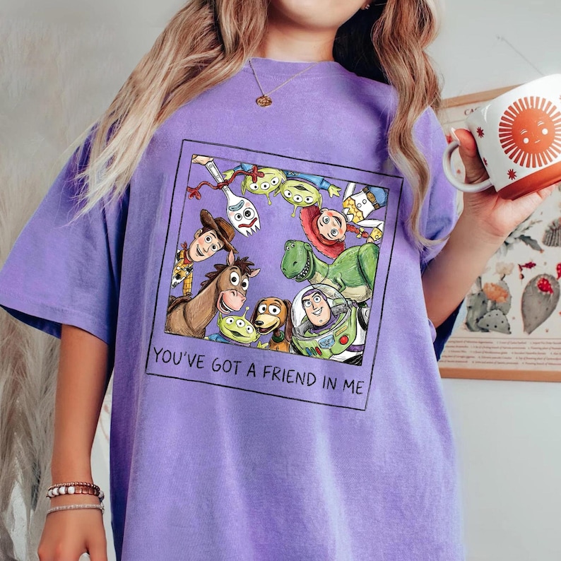 May include: A purple t-shirt featuring a framed graphic of Toy Story characters, including Woody, Buzz Lightyear, and Rex. The text "YOU'VE GOT A FRIEND IN ME" is printed below the graphic. The shirt is a solid color.