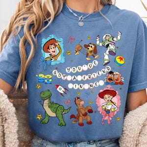 May include: A blue t-shirt featuring various Toy Story characters, including Woody, Buzz Lightyear, and Jessie. The phrase "You've Got a Friend in Me" is displayed in white letters. The design includes stars, bows, and a rocket.
