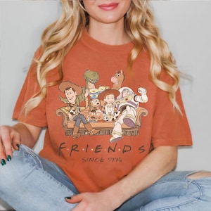 May include: Burnt orange t-shirt featuring a graphic of Toy Story characters on a couch. The word "FRIENDS" is printed below the graphic, with "SINCE 1995" underneath. The shirt is made of a soft material.